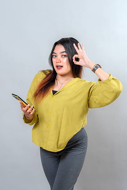 istock Confident young Asian woman smiling with a bright expression, holding a smartphone and making an OK hand gesture. Her face shows approval, positivity, and cheerful charm. 2245298209