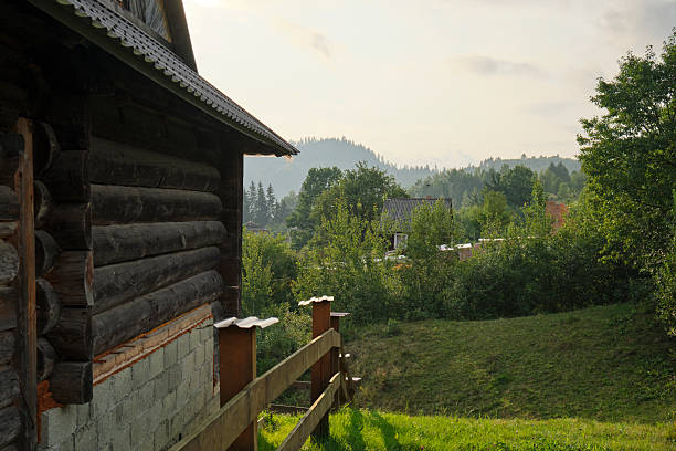 A rustic wooden cabin with a metal roof stands beside a grassy slope and fence, surrounded by lush greenery and distant forested hills. The soft natural light and tranquil setting evoke the calm of countryside life