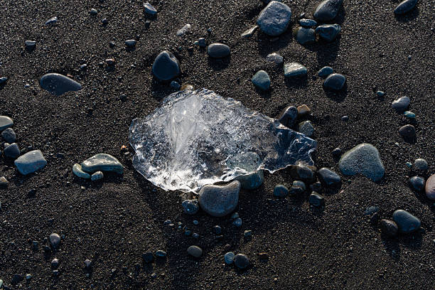 A shard of ice rests on black sand among smooth stones. The striking contrast creates texture and depth.