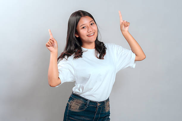 istock Cheerful young Asian woman smiling brightly while pointing both index fingers upward, showing excitement and positivity. Studio portrait with gray background. 2245301567