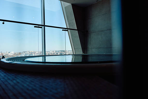 Luxury spa interior featuring a circular hot tub tub with still water reflecting sunlight, set against a large glass window overseeing a distant city landscape