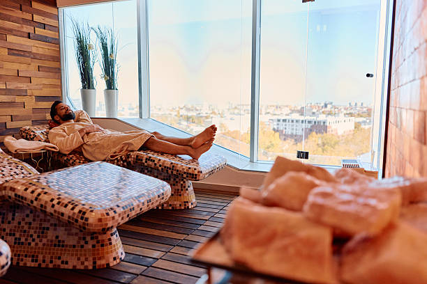 Man in bathrobe relaxing barefoot on mosaic tiled lounger in a salt room, overlooking a panoramic city skyline through large windows, serene spa wellbeing moment
