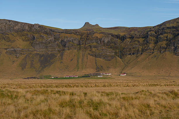 A scenic view of an Icelandic landscape with grassy fields, a mountain range, and a few houses in the valley.