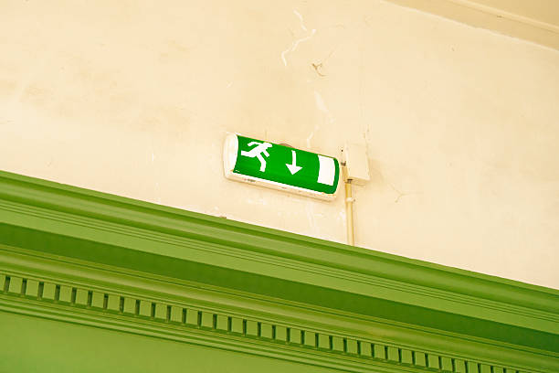 A bright green exit sign with a running figure and downward arrow is mounted on a wall. Green molding below.
