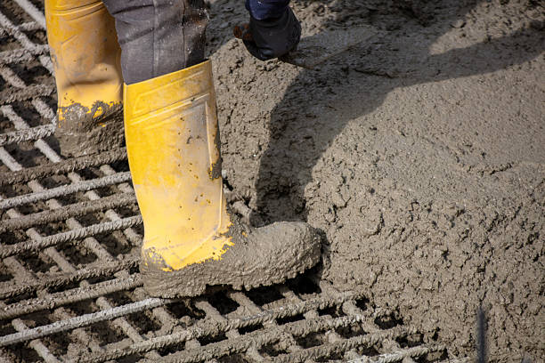 Construction worker in yellow boots spreading wet concrete over steel rebar grid at a building site under daylight.