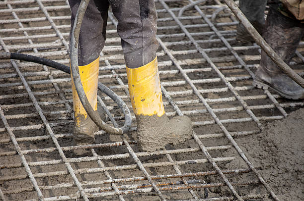 istock Worker within yellow boots pouring wet concrete over steel reinforcement grid at construction site, viewed close-up under daylight conditions 2245311701
