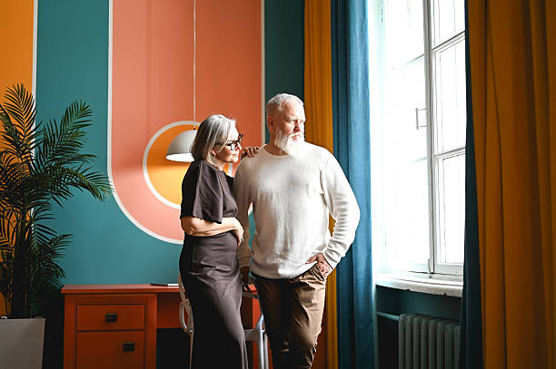 Mature couple standing by window, observing outside, enjoying togetherness