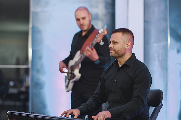 A focused keyboard player sits center stage, fingers moving across the keys as a bassist plays in the softly lit background, capturing a live music moment with a calm, professional vibe.