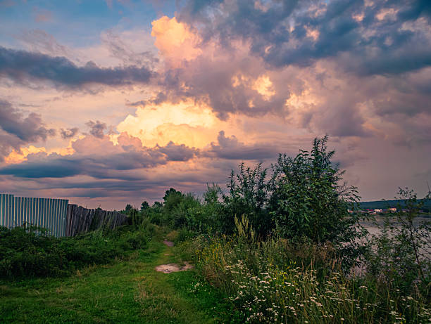 A peaceful pathway alongside the river is lined with greenery as the sun sets, casting colorful hues across the sky, highlighting the dramatic cloud formations.