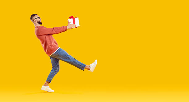 istock Funny man holding a present and walking against a yellow copy space studio background 2245325361
