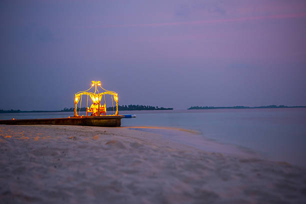 Maldives jetty pier during dusk night fall at the beach with wood walkway and palm trees during calm evening with illuminated lights