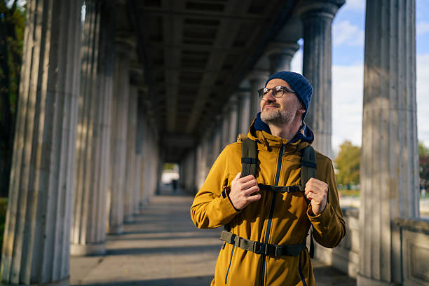 A mature man wearing a jacket and backpack stands under the famous colonnades in Berlin, enjoying the morning light. The photo captures a peaceful travel moment, curiosity, and appreciation for historical architecture in a European city.