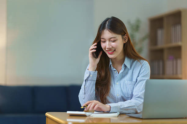 istock Professional Asian female freelancer calculating finances, talking on phone, working remotely from modern home office, displaying confident demeanor while multitasking 2245331684