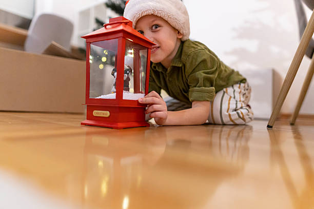 A cheerful child in a Santa hat presents a red lantern featuring a Santa Claus figure, sitting near a decorated Christmas tree with glowing lights. The bright indoor setting reflects joyful holiday spirit and family traditions during Christmas time.