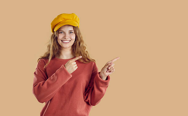 istock Young smiling woman wearing red sweatshirt and hat is pointing on copy space on beige background. 2245334779