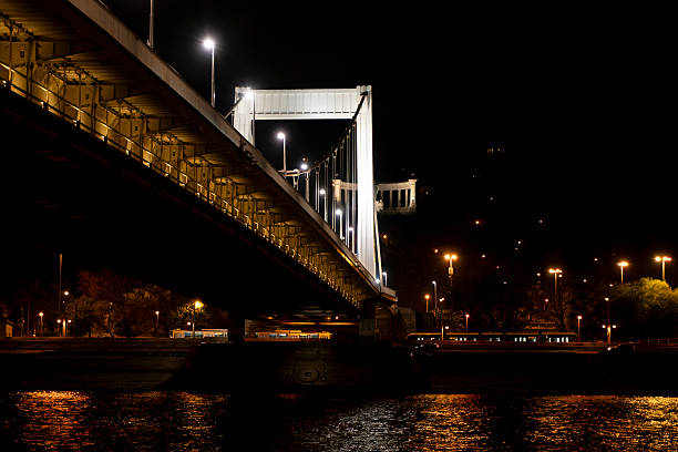 Budapest, HungaryThe iconic Széchenyi Chain Bridge stretches across the dark waters of the Danube River under a night sky in Budapest, Hungary. Suspension cables and towers glow with warm yellow lights, casting shimmering reflections on the river surface. On the Buda side, a historic castle atop Gellért Hill is visible, surrounded by scattered street lamps and illuminated paths winding up the hillside. The scene captures an urban nightscape with no people present, emphasizing architectural grandeur and serene tranquility. This image suits commercial uses such as travel brochures promoting European destinations, articles on Hungarian landmarks, city tourism campaigns, or backgrounds for media content highlighting nighttime urban beauty and infrastructure.