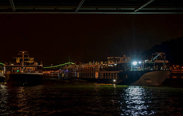 Budapest, HungaryA large white cruise ship with multiple decks is docked along a dark riverbank at night, its windows and decks glowing with warm yellow interior lights. The vessel features a sleek design with balconies and an upper lounge area visible. In the background, a prominent bridge arches over the river, illuminated by vibrant green lights along its cables, suggesting a suspension or cable-stayed structure. City buildings line the opposite shore, dotted with orange streetlights and reflections shimmering on the calm water surface. The scene captures a serene yet lively nighttime urban waterfront, ideal for commercial uses in travel brochures, tourism websites, river cruise promotions, luxury vacation articles, and cityscape illustrations for media companies targeting adventure and leisure content.