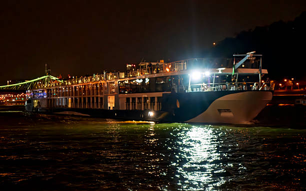 Budapest, HungaryA large white cruise ship with multiple decks is docked along a dark riverbank at night, its windows and decks glowing with warm yellow interior lights. The vessel features a sleek design with balconies and an upper lounge area visible. In the background, a prominent bridge arches over the river, illuminated by vibrant green lights along its cables, suggesting a suspension or cable-stayed structure. City buildings line the opposite shore, dotted with orange streetlights and reflections shimmering on the calm water surface. The scene captures a serene yet lively nighttime urban waterfront, ideal for commercial uses in travel brochures, tourism websites, river cruise promotions, luxury vacation articles, and cityscape illustrations for media companies targeting adventure and leisure content.