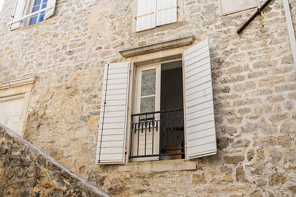 Rustic window with open wooden shutters and potted plants on stone wall. Mediterranean charm, urban gardening, and cozy architecture.
