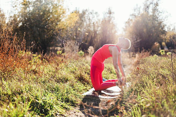 A fit woman in a red athletic outfit stretches backward in a sunlit meadow, practicing yoga outdoors. The countryside backdrop features tall grasses, warm light, and a calm, focused mood.