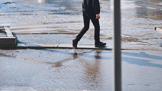 istock Young male carefully navigating flooded street, stepping on raised pavement section during rainfall aftermath, avoiding deep water puddles 2245365496
