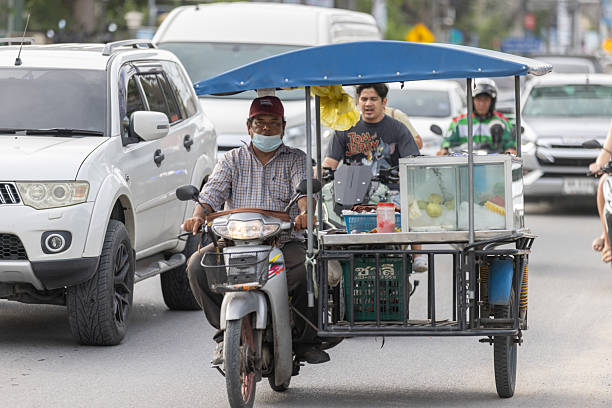06 November 2025, Ko Samui - Thailand.A typical traffic scene in Koh Samui shows a car, a motorcycle, and a tricycle-style food stall sharing the same road. The mix of regular vehicles and mobile street vendors moving side by side is a common and familiar sight in Thailand, reflecting the country’s everyday street culture.