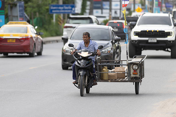 06 November 2025, Ko Samui - Thailand.A typical traffic scene in Koh Samui shows a car, a motorcycle, and a tricycle-style food stall sharing the same road. The mix of regular vehicles and mobile street vendors moving side by side is a common and familiar sight in Thailand, reflecting the country’s everyday street culture.