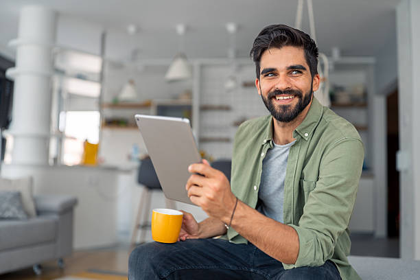 istock Smiling bearded man using tablet drinking coffee in modern bright living room with laptop nearby 2245369176