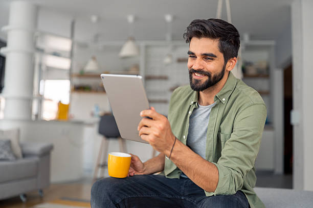istock Smiling Young Adult Male Using Digital Tablet While Drinking Coffee In Contemporary Home Interior Setting 2245369184
