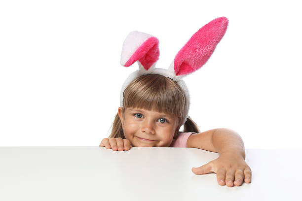 istock Curious little girl in bunny ears peeking over table against white background 2245369976