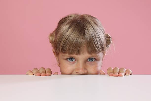 istock Curious little girl peeking over table against pink background 2245370038