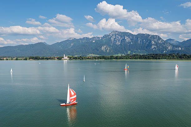 Aerial view of a summer landscape with a lake, sailboats, and mountains in the background under a blue sky on a sunny day.