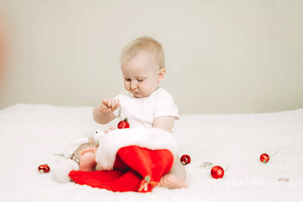 Baby sits on a white blanket, engaged with Christmas decorations while wearing a Santa hat. Cute holiday scene captures curiosity and festive spirit of the holiday season.
