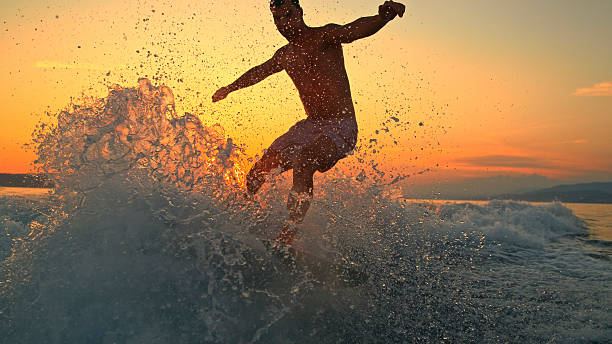 Young Caucasian male navigating waves on a surfboard behind a boat as the sun sets, creating a vibrant splash against the colorful sky.