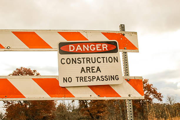 A white sign with DANGER CONSTRUCTION AREA NO TRESPASSING is mounted on a striped barrier in autumn, Infrastructure and development background or design element.