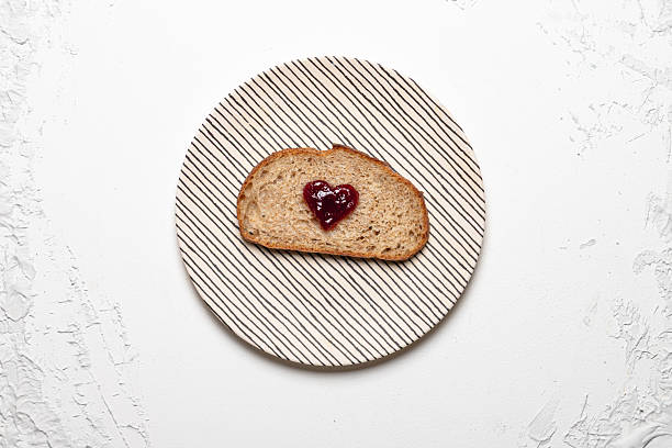 A slice of whole grain bread with a heart-shaped jam center on a striped plate over a textured white background,