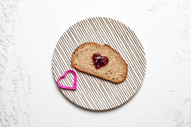 Top-down view of a slice of artisan whole-grain sourdough bread with a heart-shaped dollop of red jam on a striped plate, next to a pink heart cookie cutter, all set on a bright white textured background