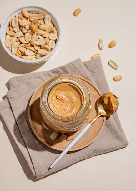 istock Peanut butter in a glass jar on a napkin on a light background with scattered nuts 2245391615