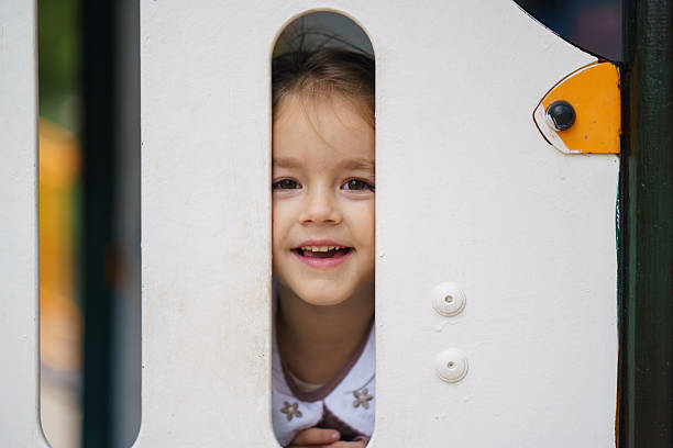 Happy girl peeking and smiling through a small opening on a playground structure.