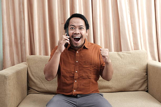 Southeast Asian man sitting on a sofa, smiling widely and cheering with excitement while talking on the phone indoors.