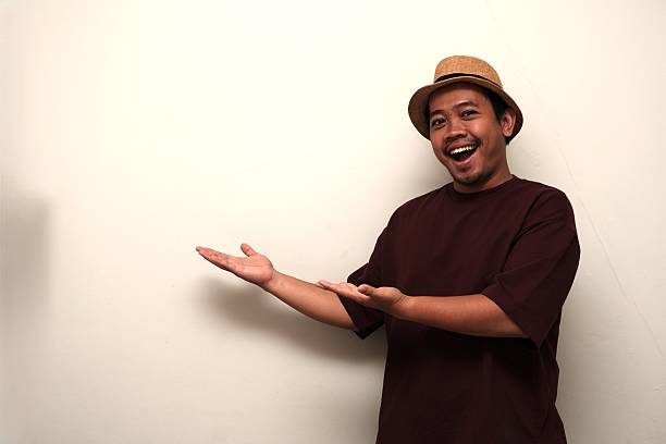 Excited Southeast Asian man in casual clothes and hat smiling while gesturing toward blank space, ideal for advertising or message.