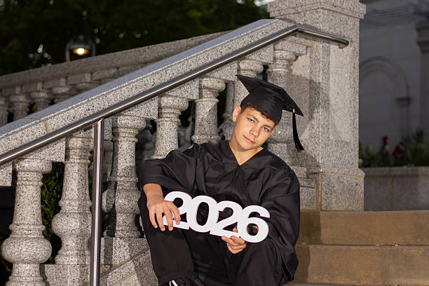 istock Young Student Celebrates Graduation While Sitting on Steps With 2026 Sign at Outdoor Ceremony During Evening Hours 2245422032