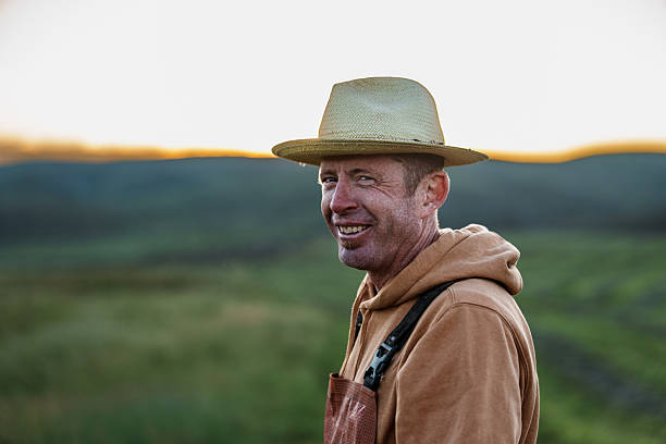A cheerful rancher wearing waterproof bibs smiles while standing in a dew-covered hay field on a peaceful Wyoming morning.