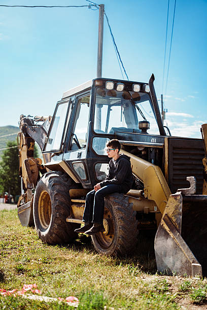A tween boy sits in the afternoon sun on the wheel of a large backhoe, taking a short break from helping his father on the family’s Wyoming ranch.