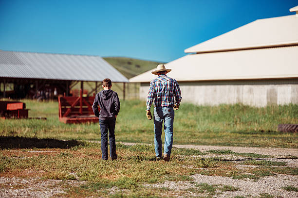 Rear view of a rancher and his tween son walking together toward the barns on their family ranch in Wyoming on a bright, sunny afternoon.