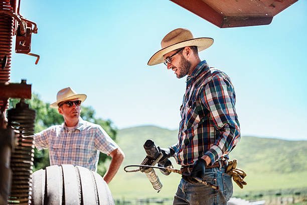 Two ranchers work together on farm machinery in the summer sun, using tools to maintain equipment for harvesting hay.