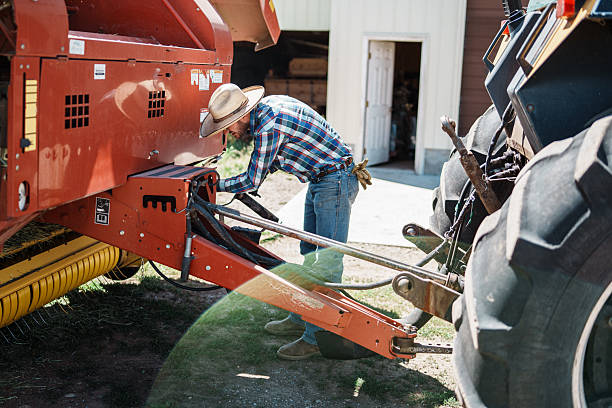 A rancher in a plaid shirt and straw hat attaches hydraulic hoses and checks connections between a tractor and hay baler before heading to the fields for hay harvest.