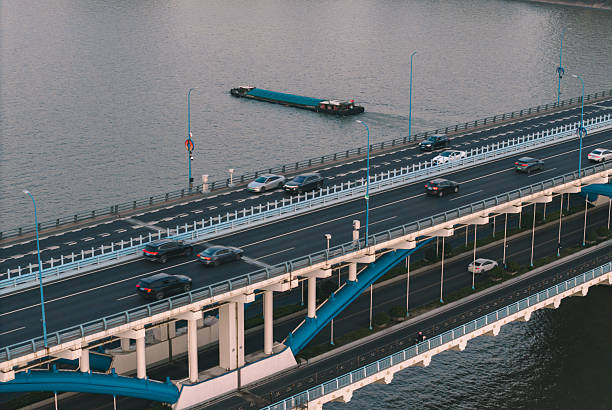 A barge moves smoothly beneath a striking blue bridge during sunset, with a vibrant city skyline rising up in the background.