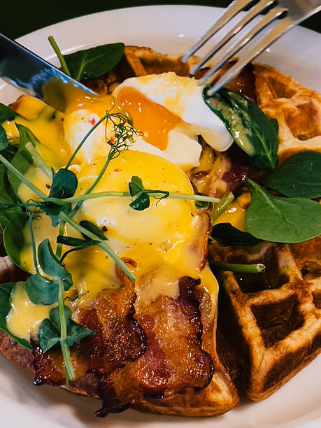 Knife and fork cutting into poached egg with runny yolk, bacon, hollandaise sauce, pea shoots and spinach on waffles, served on white plate for delicious breakfast. Gourmet healthy weekend breakfast