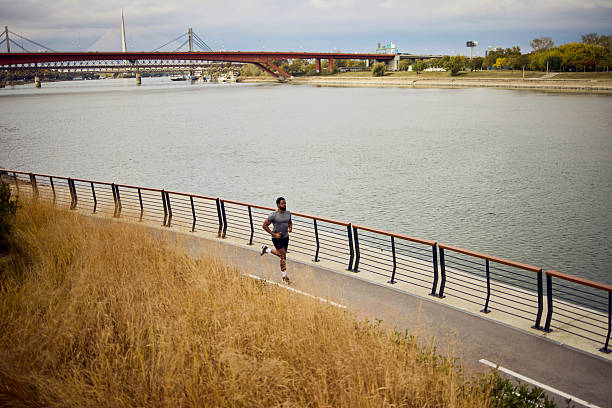 A lone runner jogs along a riverside trail beside a railing, with a distant bridge and grass in the foreground.